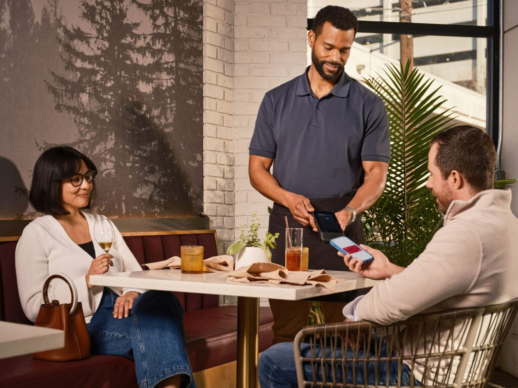 Server holding a handheld smart payment terminal at a restaurant table while a customer pays with a phone